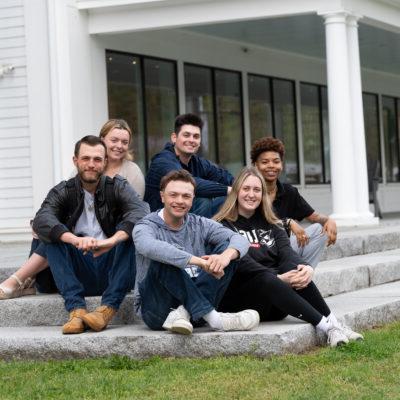 A diverse group of New England College students sit on the steps of the John Lyons Building on campus.